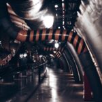 A dimly lit underground industrial corridor featuring prominent pipes and dim lighting.