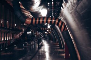 A dimly lit underground industrial corridor featuring prominent pipes and dim lighting.