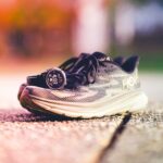 A pair of Hoka running shoes with a Garmin watch beside them on a sunny outdoor track.
