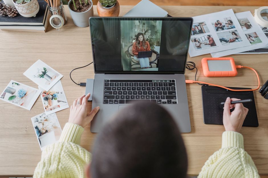 From above of crop faceless female photographer drawing on graphic tablet while editing photos on laptop at table with printed photos