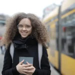 Lunettes connectées : filmer des femmes sans consentement Happy female student with curly hair in casual warm coat with backpack holding book in hands while standing on pavement on street and looking at camera