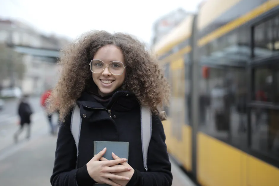 Happy female student with curly hair in casual warm coat with backpack holding book in hands while standing on pavement on street and looking at camera