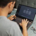 Quand l'IA conseille, la responsabilité reste humaine Man using laptop with AI interface, typing with focused attention. Indoors with eyewear beside.