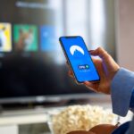 Surfshark réorganise son application iPhone et son extension Person using VPN on smartphone while watching smart TV at home.