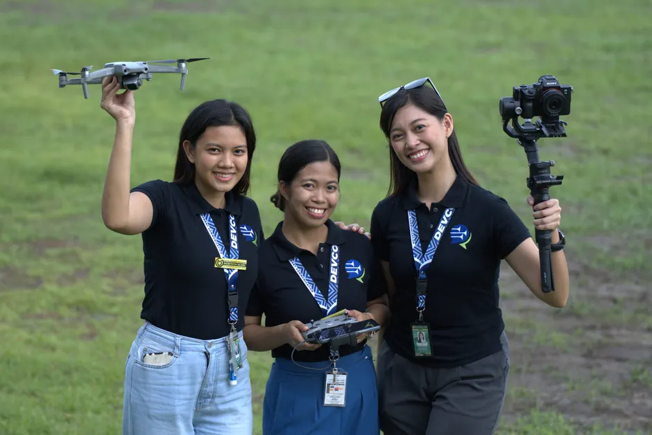 Three young females outdoors holding drone and camera equipment, smiling energetically.
