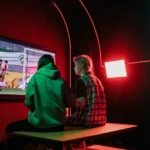 TV RGB 2026 : Sony, Samsung ou OLED selon votre usage Two friends immersed in gaming on a large screen, surrounded by vibrant RGB lighting in a cozy setup.