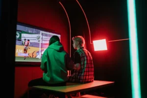Two friends immersed in gaming on a large screen, surrounded by vibrant RGB lighting in a cozy setup.
