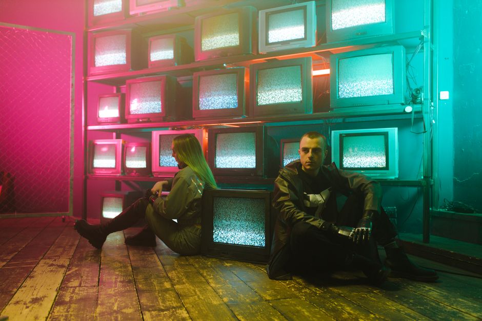 Two individuals in a cyberpunk setting with neon lighting and stacked television screens.